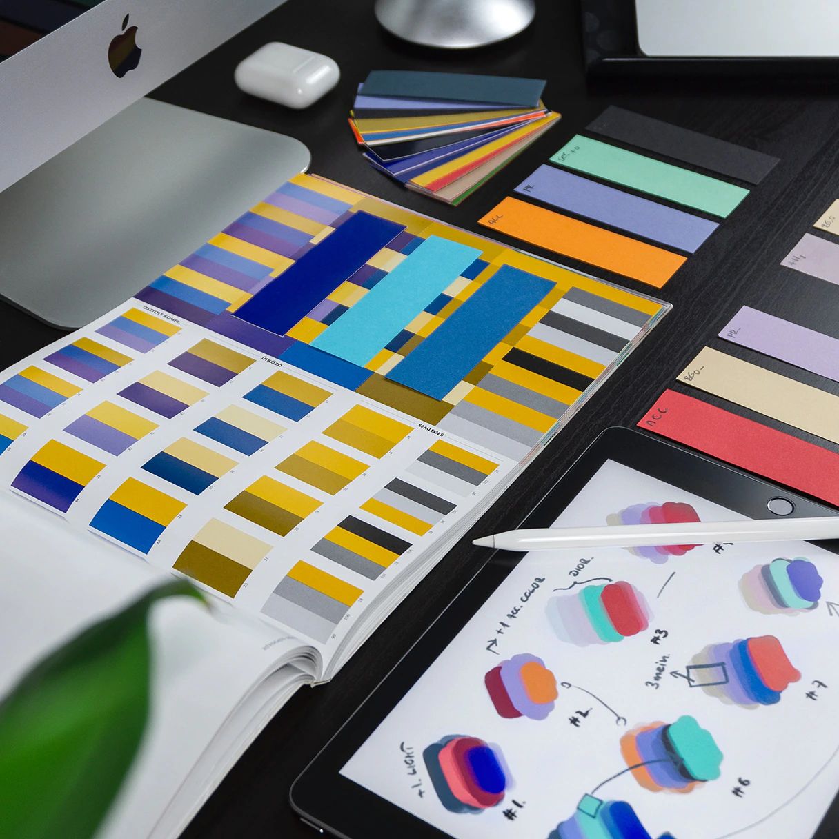 Arbeitsplatz eines Grafik Designers mit Hardware von Apple bestehend aus einem iMac und einem iPad Tablet mit iPen Stift. Auf dem Arbeitsplatz liegen Farbfächer und Broschüren.