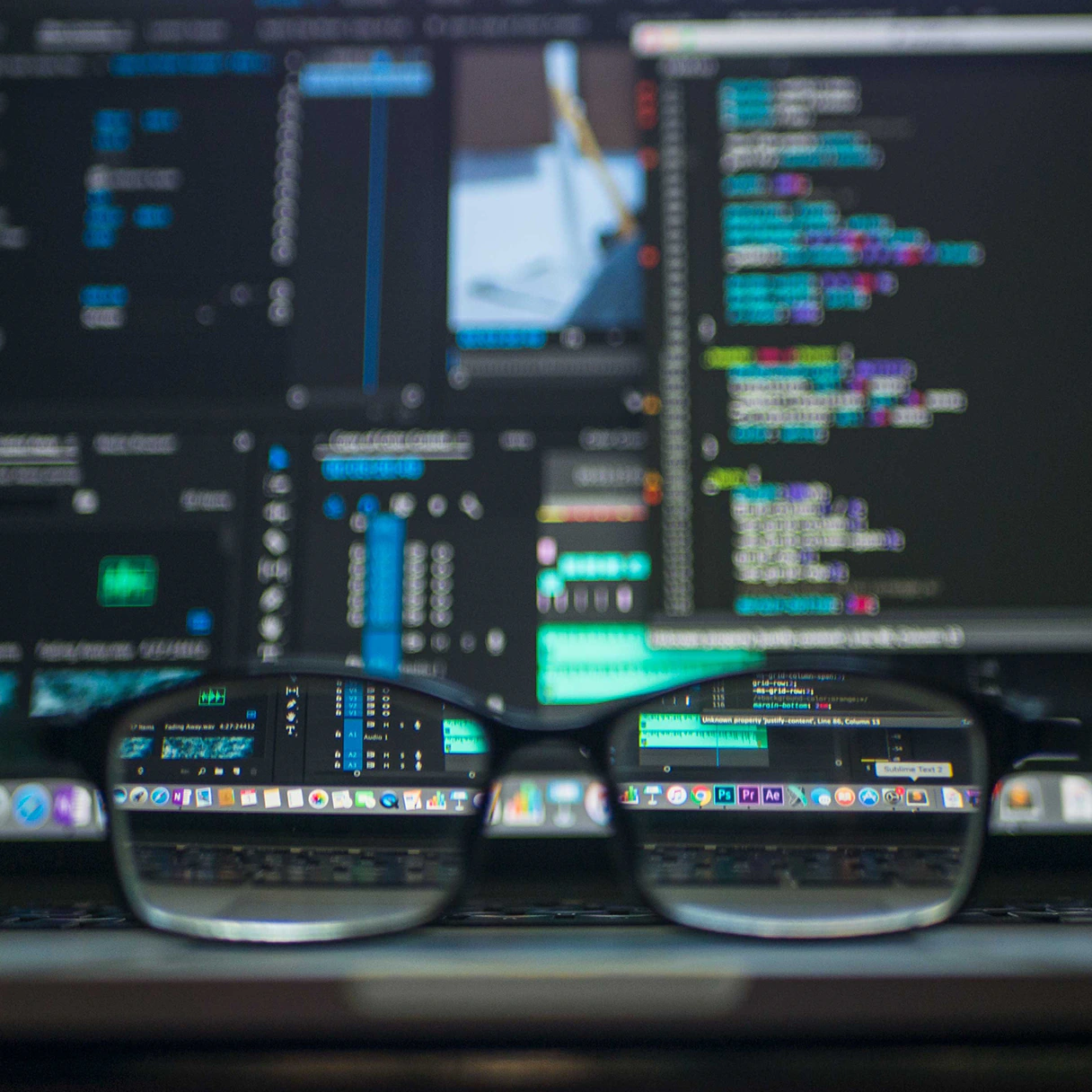 Schreibtisch eines Programmierers auf dem eine Brille liegt. Der Hintergrund ist unscharf, es ist aber Programmierungs-Code zu erkennen.